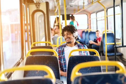 Handsome young smiling African American guy sitting in public transportation and listening to the music while looking through the window.