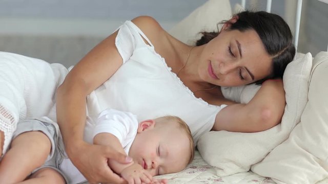 Happy mom is lying with her newborn sleeping baby in bed. Mom sleeps next to the baby.