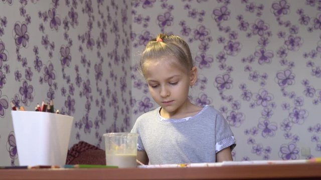 A little cute girl draws colored paints on a sheet of paper in her room