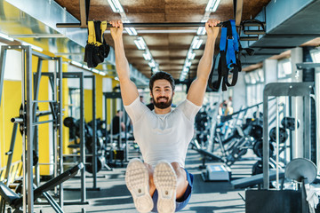 Smiling bearded muscular man in sportswear hanging from the bar and doing endurance. Gym interior.