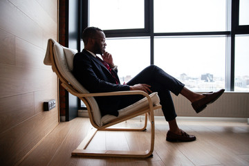 Thoughtful African businessman in a suit sitting in a chair on the background of a panoramic window...