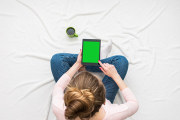 Woman woman uses a tablet, sitting on a white crumpled blanket, green Cup of coffee or tea, women's feet. Women's hands. Background with copy space, for advertisement. Top view