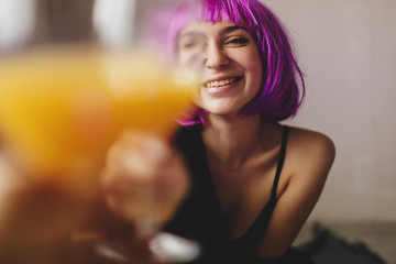 Attractive playfull woman wears pink wig, black lingerie and shirt. Girl drinking orange juice in glass. Woman sitting on the floor. She look sexy and hot, girl raised a glass. Toast