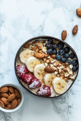 Healthy breakfast berry smoothie coconut bowl topped with blueberry,almond, banana, granola and raspberries. Top view. white marble background.