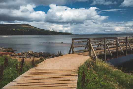 Close-up wooden pier connecting the sea and grass covered land. Northern Ireland. Stunning shoreline among the wild virgin nature. Blue cloudy sky background. Amazing Irish landscape.
