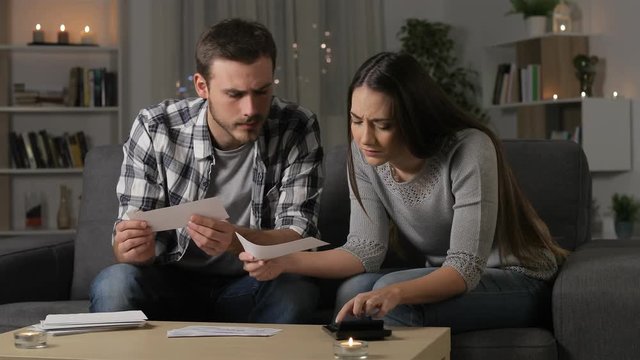 Sad couple checking bank receipts sitting on a couch in the night at home