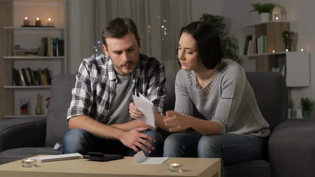 Sad couple complaining reading a letter in the night sitting on a couch at home