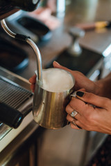 Fototapeta na wymiar Female hands holding silver pitcher near coffee machine, barista steaming milk before adding it to espresso to make cappuccino or latte