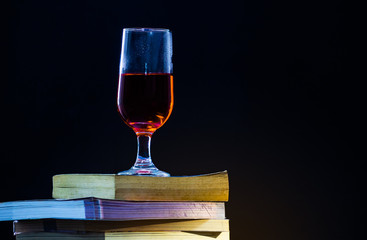 Old books overlap In a black background and with dim light one glass of red wine on top.
