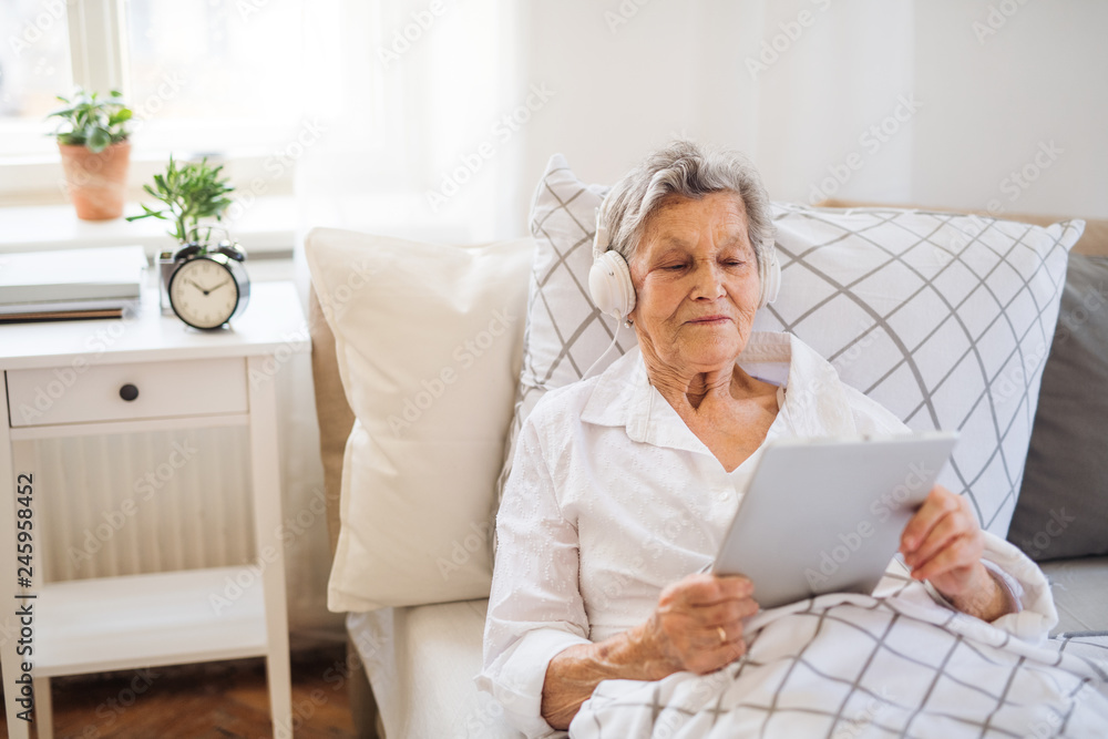 Wall mural Sick senior woman with headphones and tablet lying in bed at home or in hospital.