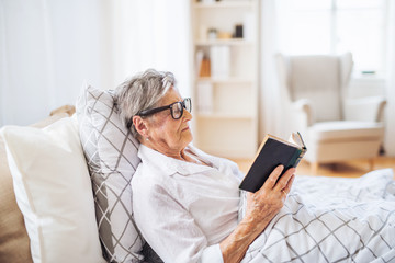 Sick senior woman reading bible book in bed at home or in hospital.
