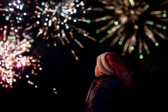 Side view of a boy wearing a santa hat standing outdoors watching a Christmas firework display at night