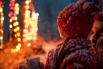 Close-up of a girl standing outdoors looking at illuminated Christmas lights holding a hot chocolate drink, USA