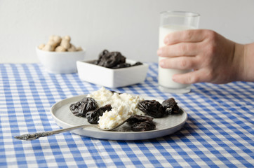 Healthy delicious breakfast time. Teenage hand taking glass of milk. Plate with cottage cheese, prunes, peanuts on table. Served table with blue  checkered tablecloth