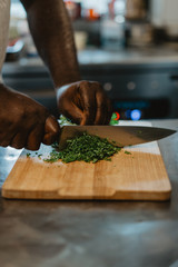 Black chef cutting greens on the kitchen of restaurant. Close up image of dark skin african cook preparing dishes in a healthy food hipster cafe. Concept of vegetarian lifestyle
