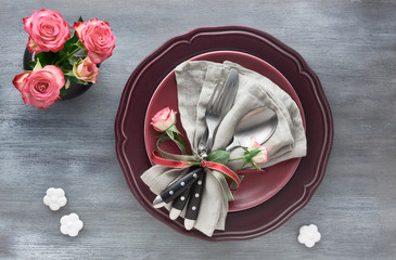 Valentine's day, birthday or anniversary table setup, top view on greybackground. Pink roses, dark red plates, napkin and crockery, decorated with rose bud and ribbons.