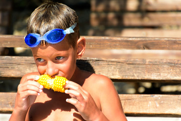 Happy boy wearing swimming goggles eating corn on the cob. Summer weekend
