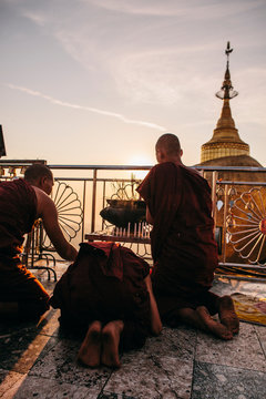 Monks in maroon robes kneel at sunset to light candles and incense in front of giant gold-leaf gilded rock and stupa.