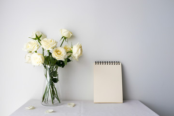 White roses in a glass vase and white blank notepad on white table top on white background with copy space for product mockup placement for valentine's day wedding ceremony