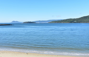 Beach in a bay with island, small coastal village and forest. Bright sand and blue sea with foam, sunny day, clear sky. Galicia, Spain.
