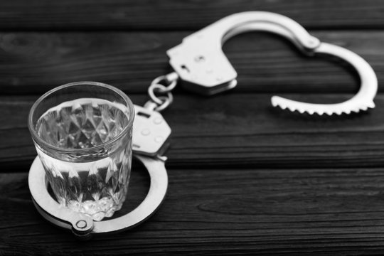 Iron handcuffs for the arrest of criminals, a glass of vodka on a wooden. background. a crime committed while intoxicated.