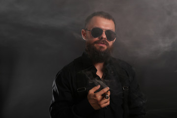 young handsome man with big beard and black sunglasses on black background  smoking cigar 