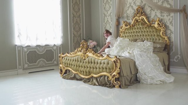 Young laughing beautiful woman in white vintage ball costume running and jumping to the huge golden bed