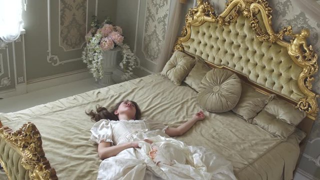 Young woman in ball gown falls on the gold decorated bed holding silk hat. Girl is tired and sleepy