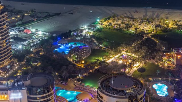 Aerial view of modern illuminated skyscrapers and beach at Jumeirah Beach Residence (JBR) night timelapse in Dubai, UAE. Green lawn and pools
