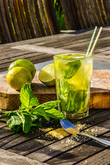 Cold refreshing summer alcoholic or non-alcoholic drink Mojito cocktail in glass decorated with fresh mint, limes and sugar. Ingredients on wooden table in the farden outdoor.