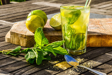 Cold refreshing summer alcoholic or non-alcoholic drink Mojito cocktail in glass decorated with fresh mint, limes and sugar. Ingredients on wooden table in the farden outdoor.