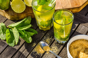 Cold refreshing summer alcoholic or non-alcoholic drink Mojito cocktail in glass decorated with fresh mint, limes and sugar. Ingredients on wooden table in the farden outdoor.