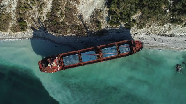 Aerial top view of an empty, red barge moored near the beautiful blue sea shore. Big Industrial ship standing near the hill with green trees.