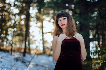 Portrait of a beautiful girl in a dark long dress who stands alone among the trees in the winter forest