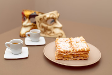 Typical Italian carnival fritters (Chiacchiere di Carnevale) dusted with powdered. Composition with two cups of coffee and in the background are Venetian carnival masks. Italian Carnival sweets. 
