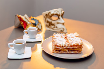 Typical Italian carnival fritters (Chiacchiere di Carnevale) dusted with powdered. Composition with two cups of coffee and in the background are Venetian carnival masks. Italian Carnival sweets.