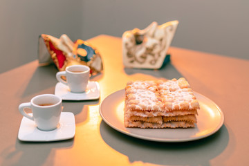 Typical Italian carnival fritters (Chiacchiere di Carnevale) dusted with powdered. Composition with two cups of coffee and in the background are Venetian carnival masks. Italian Carnival sweets.