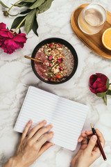Flatlay of woman's hands with notebook and smartphone, vegan smoothie bowl with chia pudding topped with granola, kiwi, pine nuts and rose buds and peon flowers on marble, healthy breakfast concept