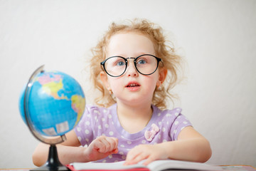 Close up photo portrait of funny little girl with glasses. She's leafing through the book on the table is a globe