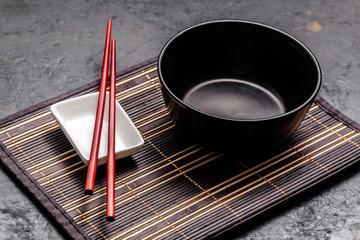 Empty Japanese dishes. A black ceramic bowl for Chinese noodles or Thai soup lies on a bamkuk rug. White saucepot for soy sauce and red Chinese sticks on a black background. Top view, copy space