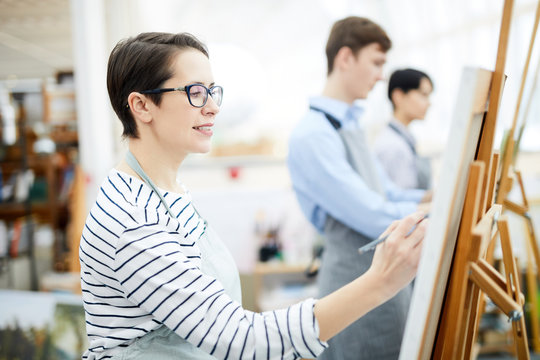 Side view portrait of smiling adult woman painting on easel enjoying work in art class, copy space