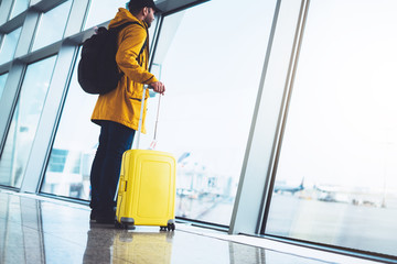 tourist with yellow suitcase backpack is standing at airport on background large window, traveler person waiting in departure lounge area hall of ​​the airport lobby terminal, vacation trip concept