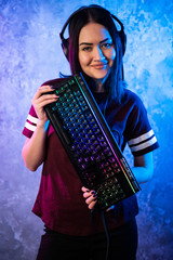 Beautiful Friendly Pro Gamer Streamer Girl Posing With a Keyboard in Her Hands, Wearing Glasses. Attractive Geek Girl with Cool Neon Retro Colors in Background. © romankosolapov