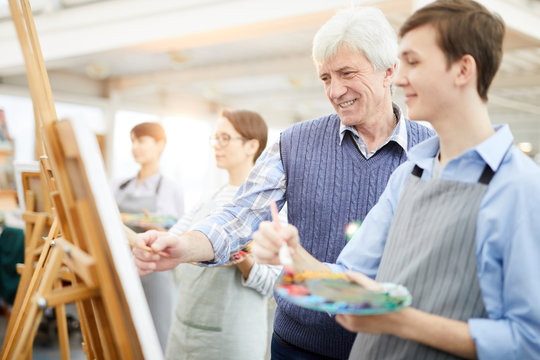 Waist up portrait of smiling art teacher helping student painting picture on easel during class, copy space