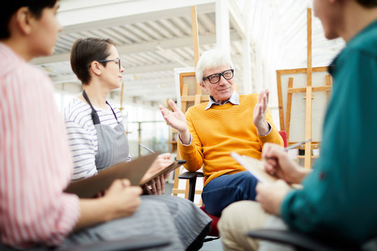 Portrait of mature art teacher talking to group of students sitting in circle in art studio