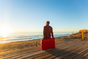Fototapeta premium Nature, travel and people concept - rear view of man sitting on red suitcase near the sea and watching to the sunset