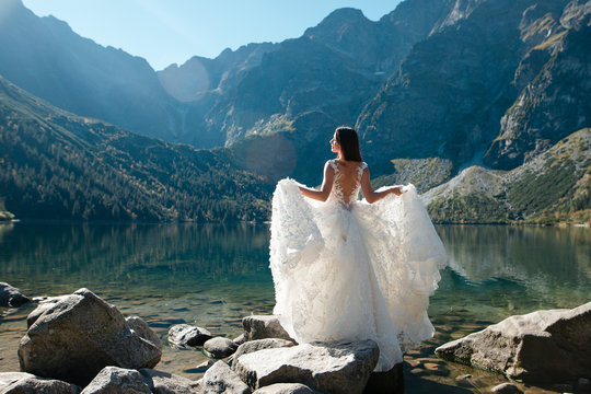 Back view of bride in beautiful wedding dress standing on the lake shore with scenic mountain view