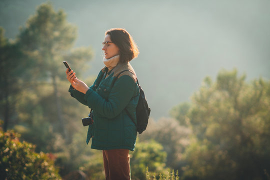 Young hipster female traveller with a backpack and photo camera watching the route in her mobile phone with wireless internet and sending pictures to social media, forest and nature in the background