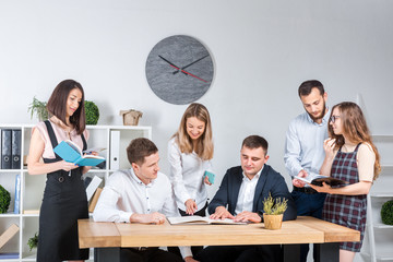 Theme is business and teamwork. A group of young Caucasian people office workers holding a meeting, briefing, working with papers and documents in a light office office around a wooden table