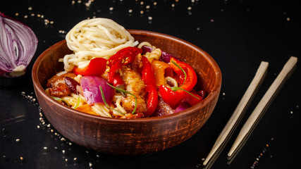 Fototapeta na wymiar Japanese, Chinese udon noodles with chicken and vegetables, Bulgarian pepper, red onion, white and black sesame. dish in a ceramic dish made of red clay on a black background.